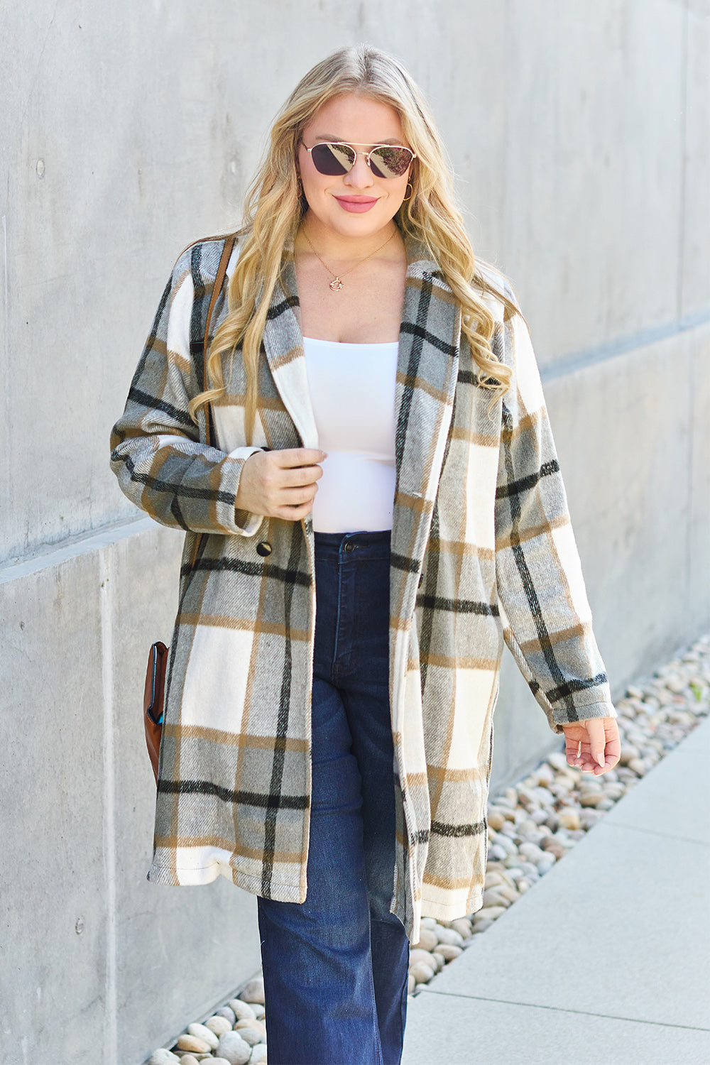 A woman stands on a sidewalk wearing the Double Take Full Size Plaid Button Up Lapel Collar Coat in pink and white over a white top and blue jeans, with her right hand lightly touching her hair. The polyester fabric of the coat makes it durable and perfect for an easy machine wash cold.