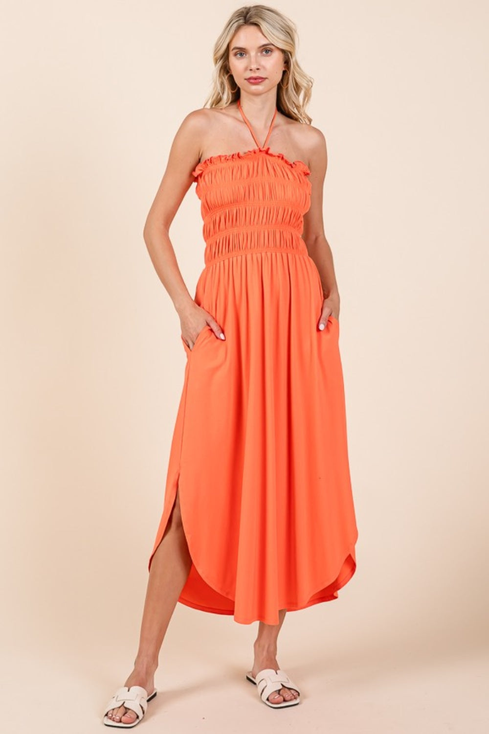 A woman wearing the Culture Code Tie Back Shirring Dress with Pockets, a versatile wardrobe staple in orange, stands against a plain background, smiling with her hands comfortably resting in the pockets.