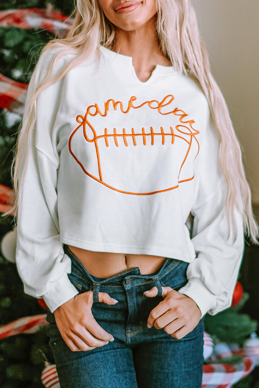 A woman with long blonde hair dressed in a chic White Game Day Lettering Rugby Football Notched Neck Sweatshirt and blue jeans stands near a Christmas tree adorned with red and white ornaments and ribbon.