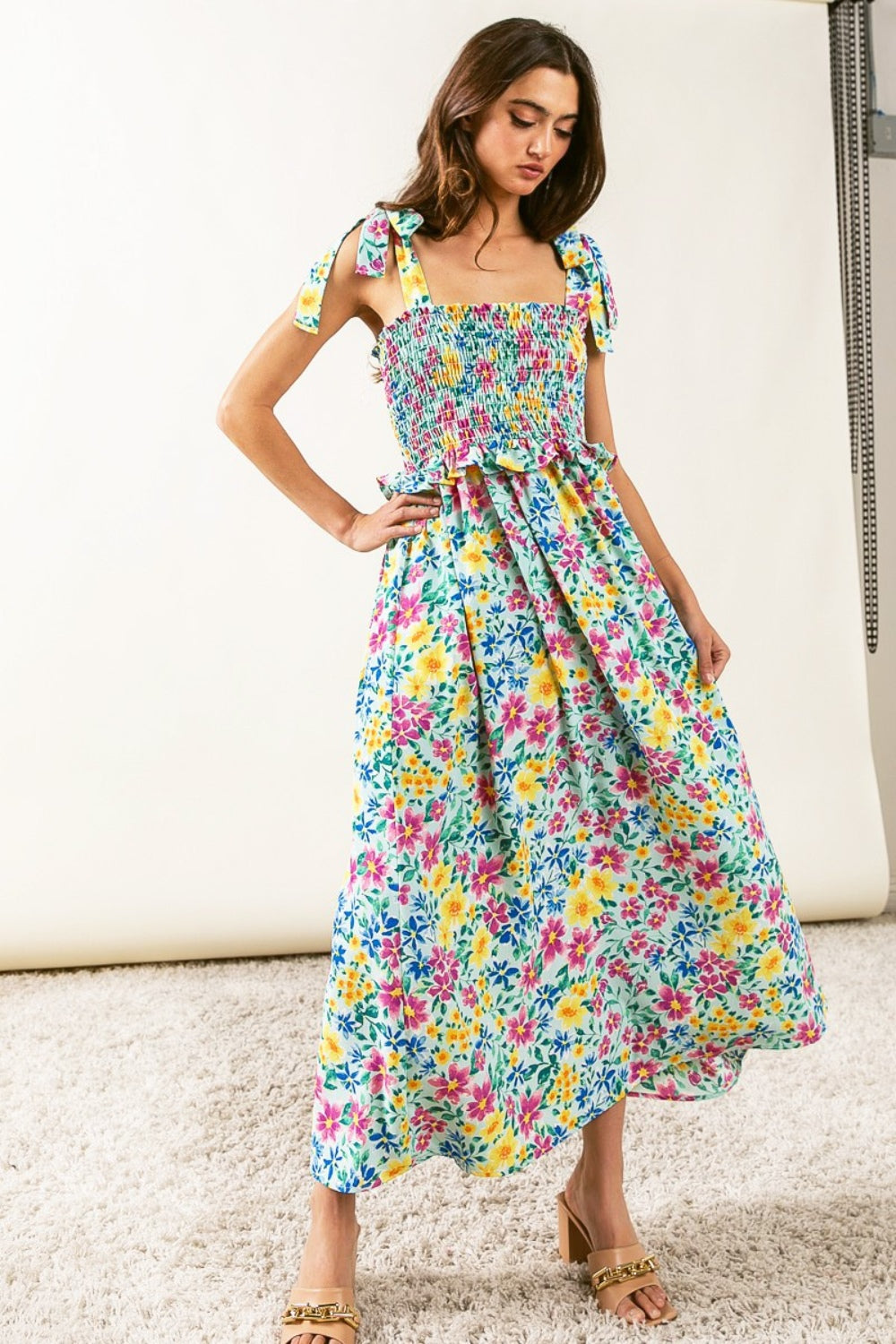 A person wearing the BiBi Floral Ruffle Trim Smocked Cami Dress with beige sandals stands on a beige carpet, posing against a white backdrop.