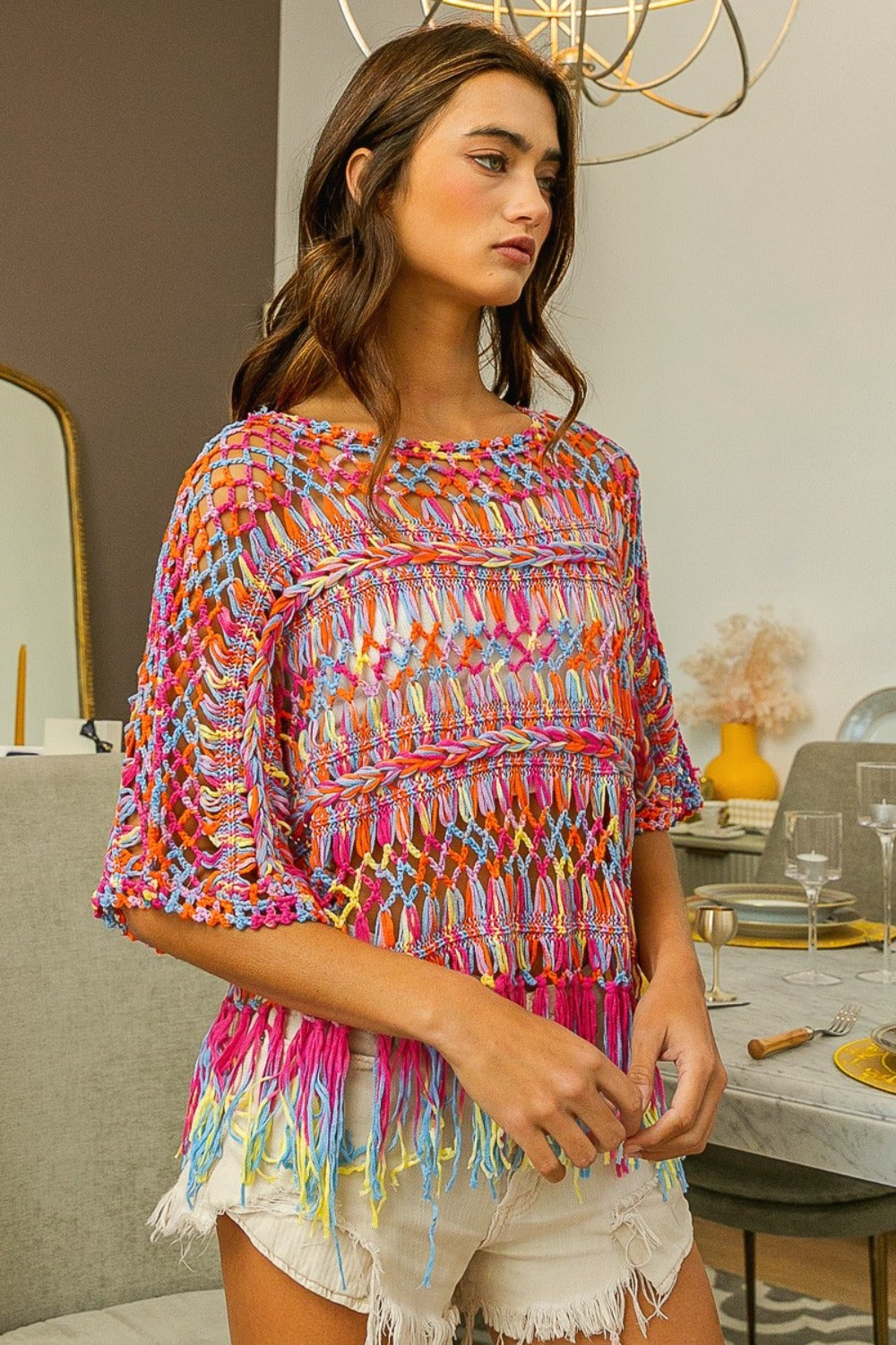 A woman stands indoors wearing the BiBi Openwork Fringed Knit Cover Up over a white outfit, radiating bohemian flair. This versatile piece adds vibrancy to her summer wardrobe.