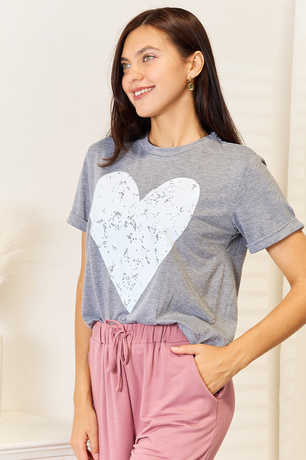 A woman with long hair stands indoors, smiling and touching her hair, wearing the trendy Simply Love Heart Graphic Cuffed Short Sleeve T-Shirt in gray.