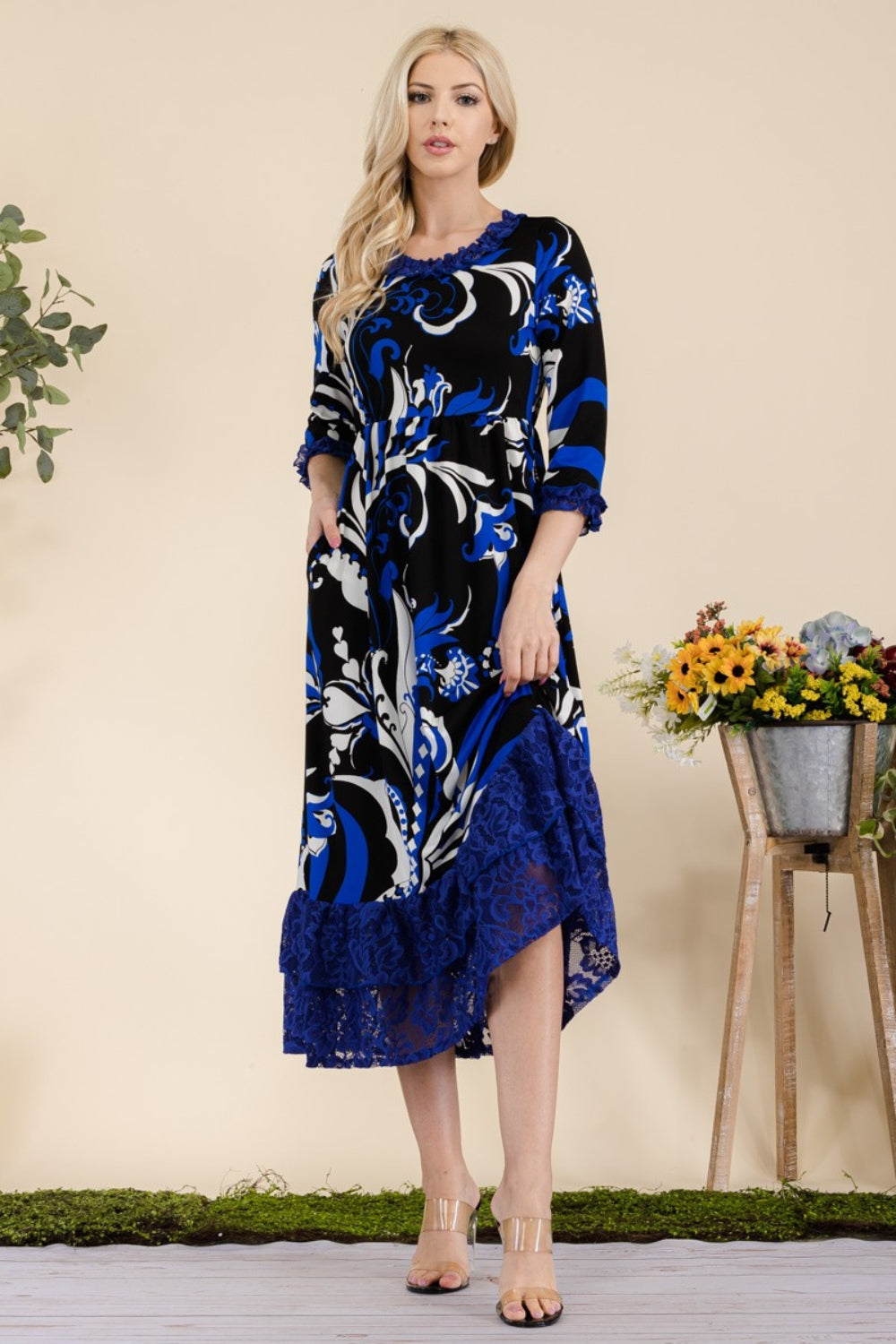 A woman wearing the Celeste Full Size Paisley Print Lace Ruffled Midi Dress stands near a wooden table adorned with a bucket of flowers.