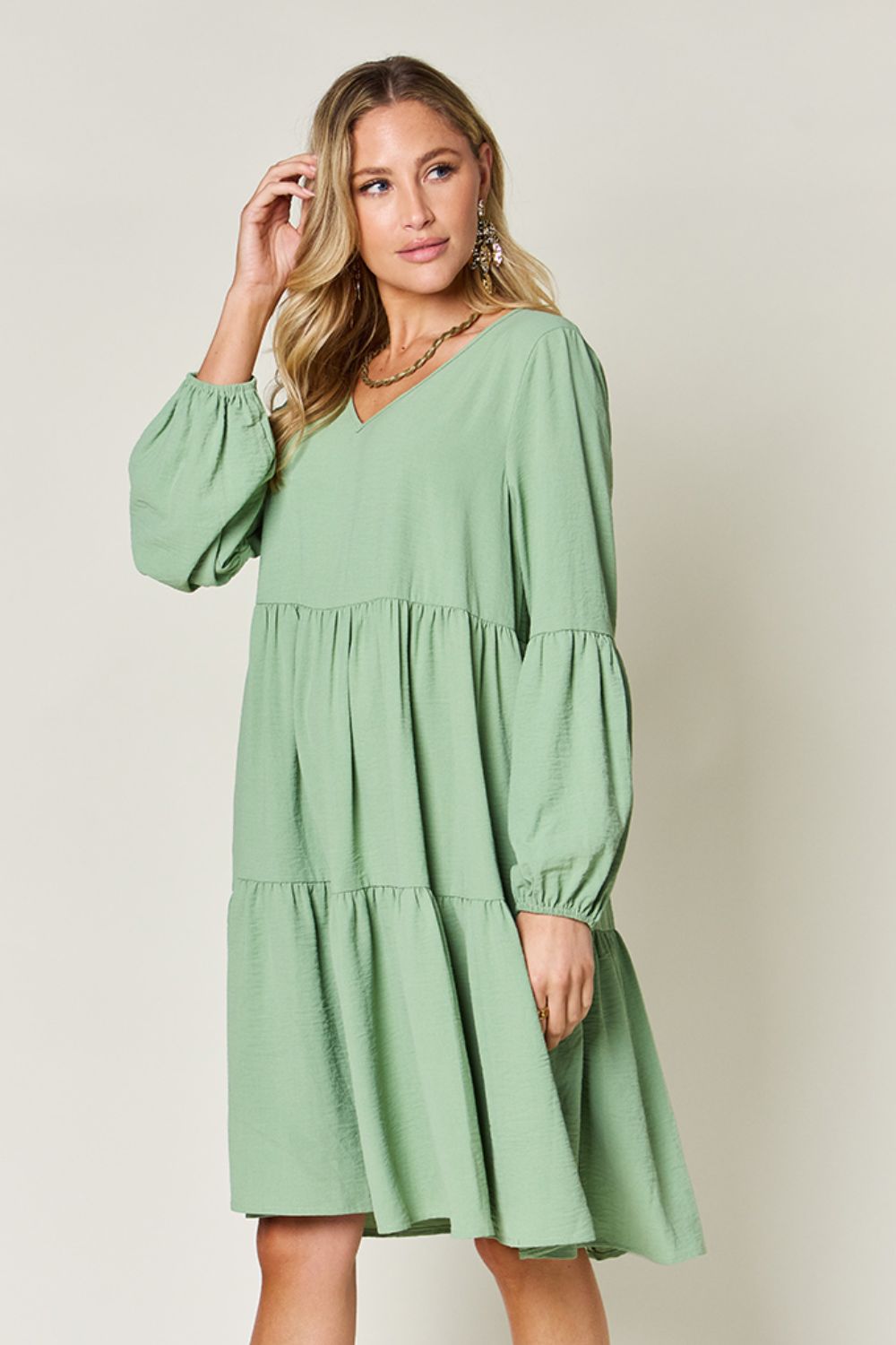 A woman stands wearing the Double Take Full Size V-Neck Balloon Sleeve Tiered Dress with Pockets in pink. Crafted from high stretch yarn, this dress combines comfort and style effortlessly. She has long, wavy hair and accessorizes with hoop earrings and a necklace. This imported garment is easy to care for – simply machine wash cold to keep it looking fresh.