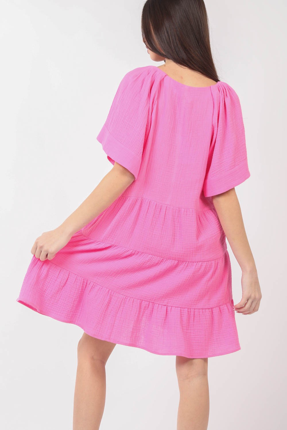 A woman is posing against a plain background, wearing the VERY J Texture V-Neck Ruffled Tiered Dress in pink, featuring short sleeves.