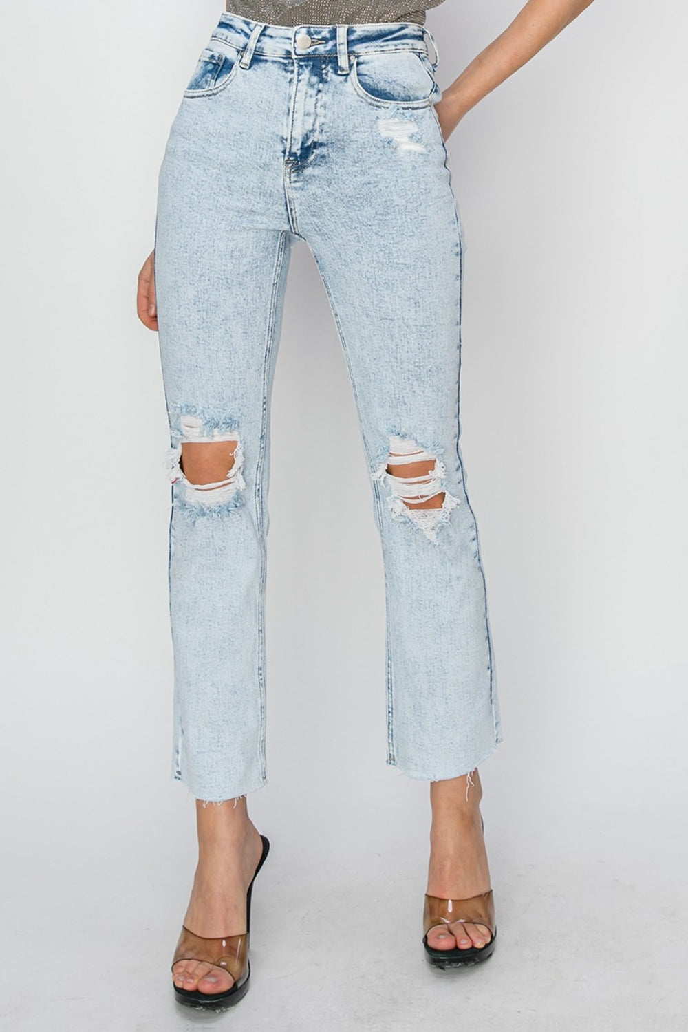 Person wearing RISEN High Rise Distressed Ankle Jeans in light blue with rips and raw hems, paired with transparent-strap high heels.