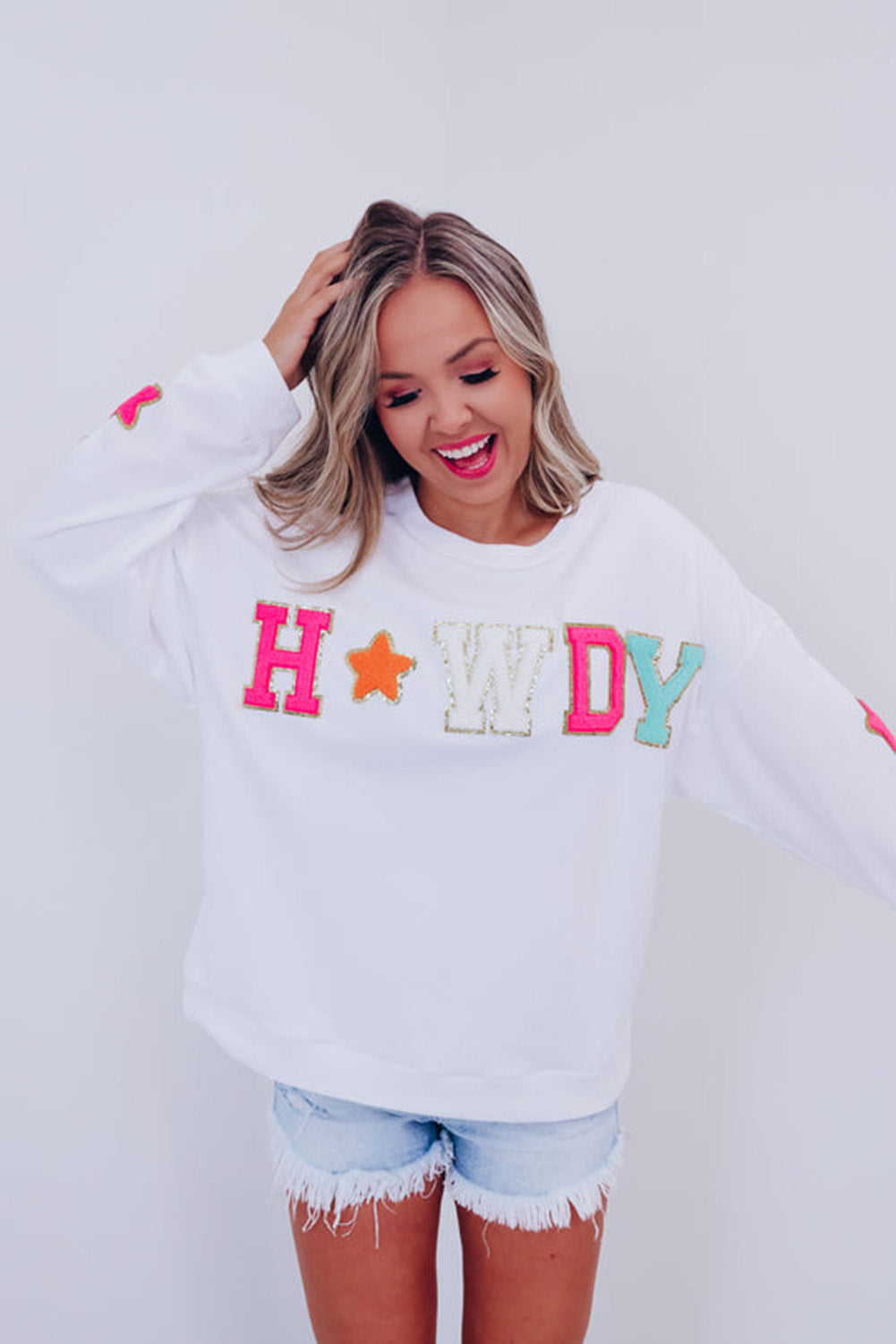 A person poses in the White Howdy Glitter Chenille Patch Graphic Casual Sweatshirt, adorned with colorful star patterns and the letters "WD." She wears light denim shorts and has shoulder-length blonde hair.