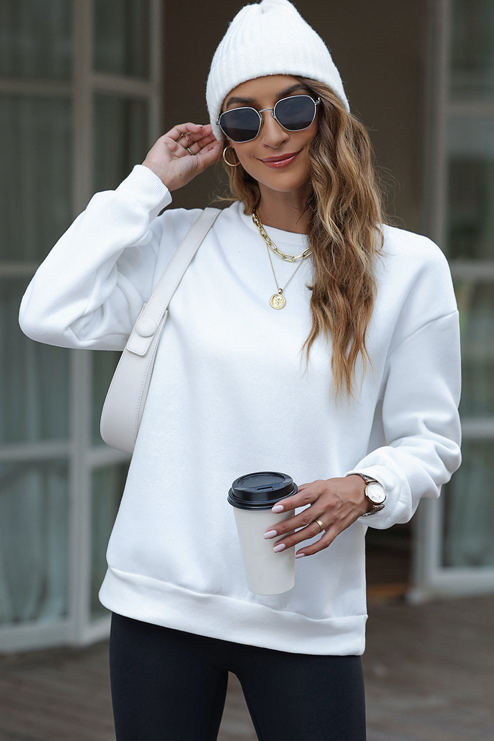 A person wearing a white beanie, the EVERY THING WILL BE OKAY Colorful Letters Sweatshirt, and sunglasses, holding a coffee cup, stands outside near windows.