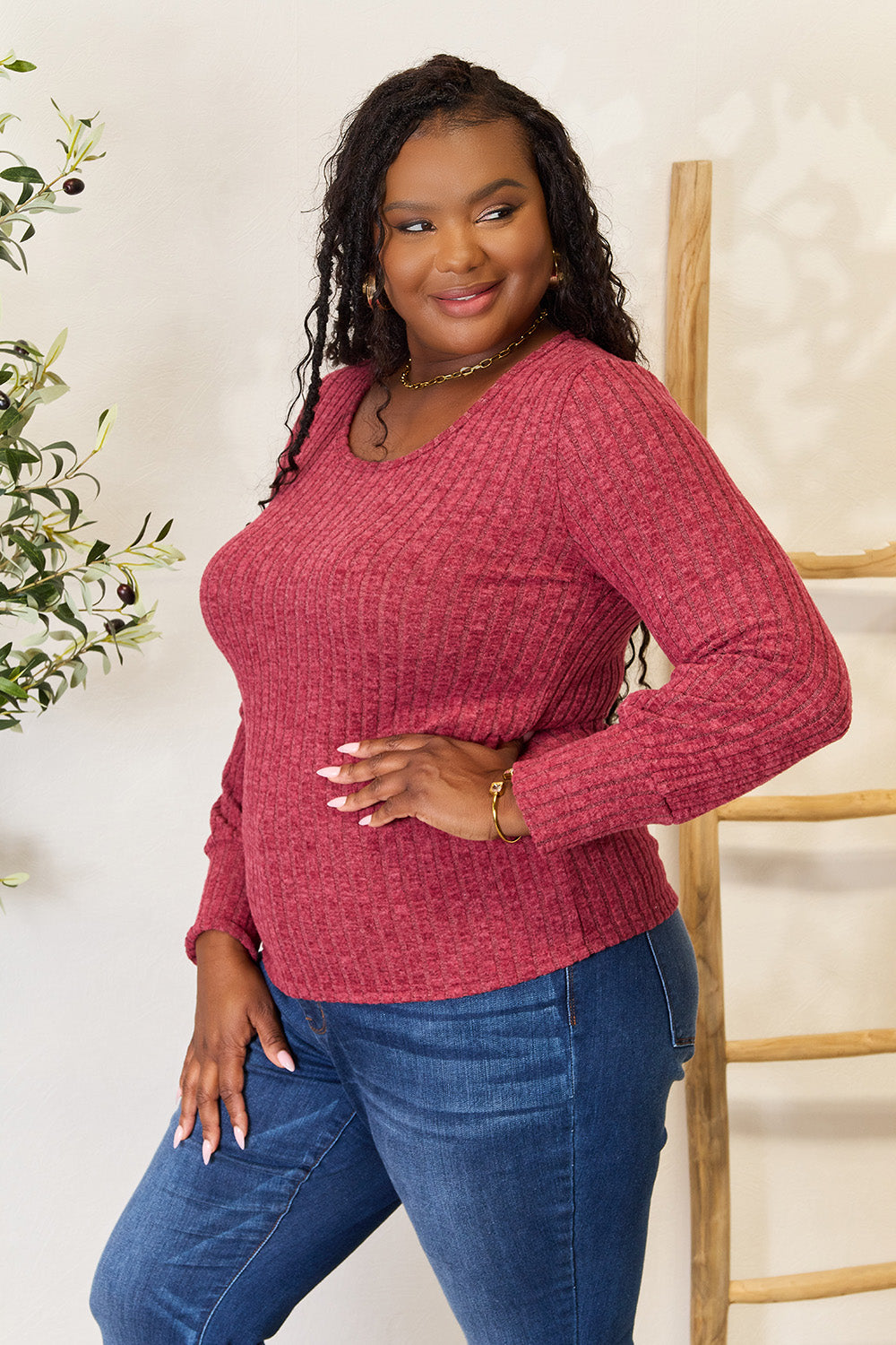 A person in a stylish maroon Ribbed Round Neck Lantern Sleeve Blouse and jeans stands by a wooden ladder, with olive branches in the background.