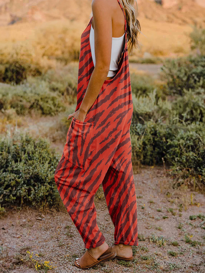 A person wearing a casual white V-neck tank top and a Full Size Printed V-Neck Sleeveless Jumpsuit with a brown animal print zebra-stripe pattern stands outdoors with one hand on their hat and the other in their pocket, set against a rocky landscape backdrop.