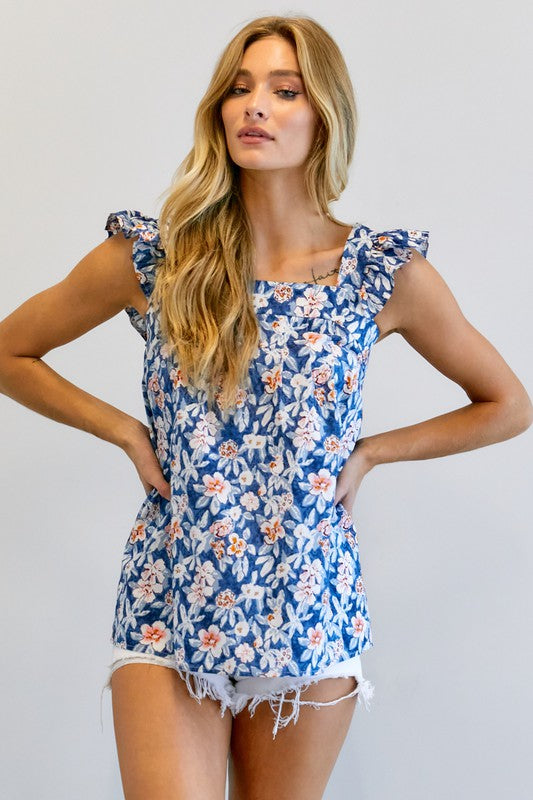 A woman in a vibrant summer wardrobe stands against a plain background, wearing the Floral Printed Ruffle Sleeveless Top paired with white shorts, with one hand casually tucked in her pocket.