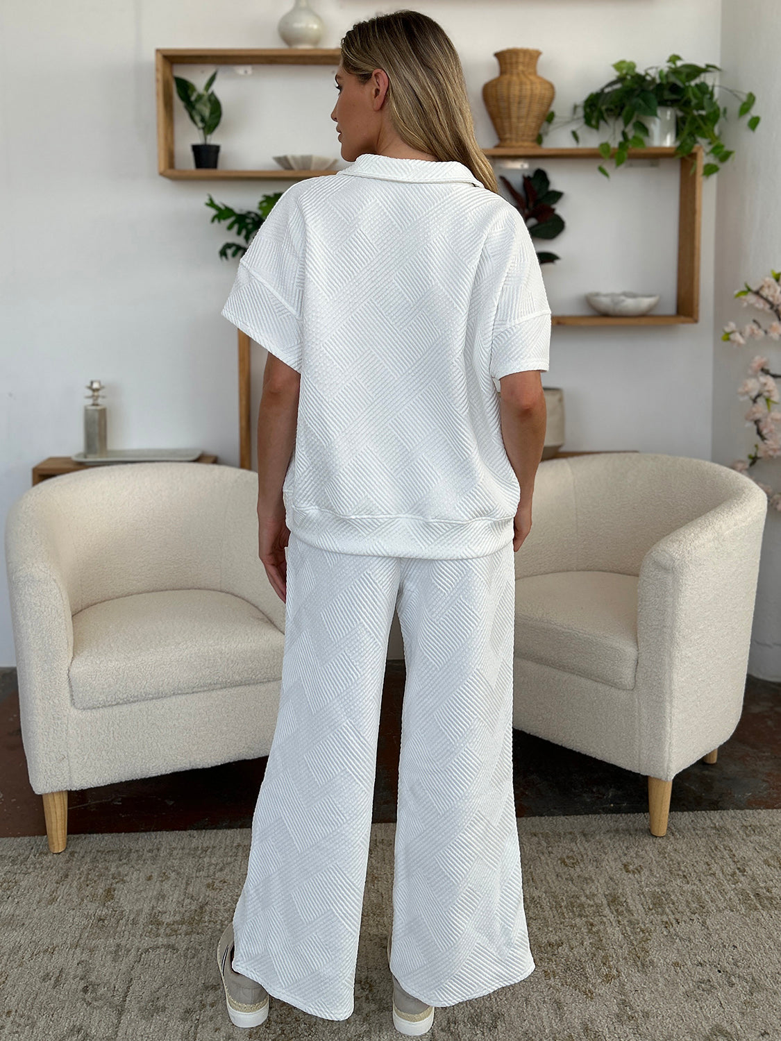 A woman is wearing the Double Take Full Size Texture Half Zip Short Sleeve Top and Pants Set, featuring a white zip-up short-sleeve top and matching wide-leg, drawstring pants. She stands with one hand in her pocket and smiles against a light background.