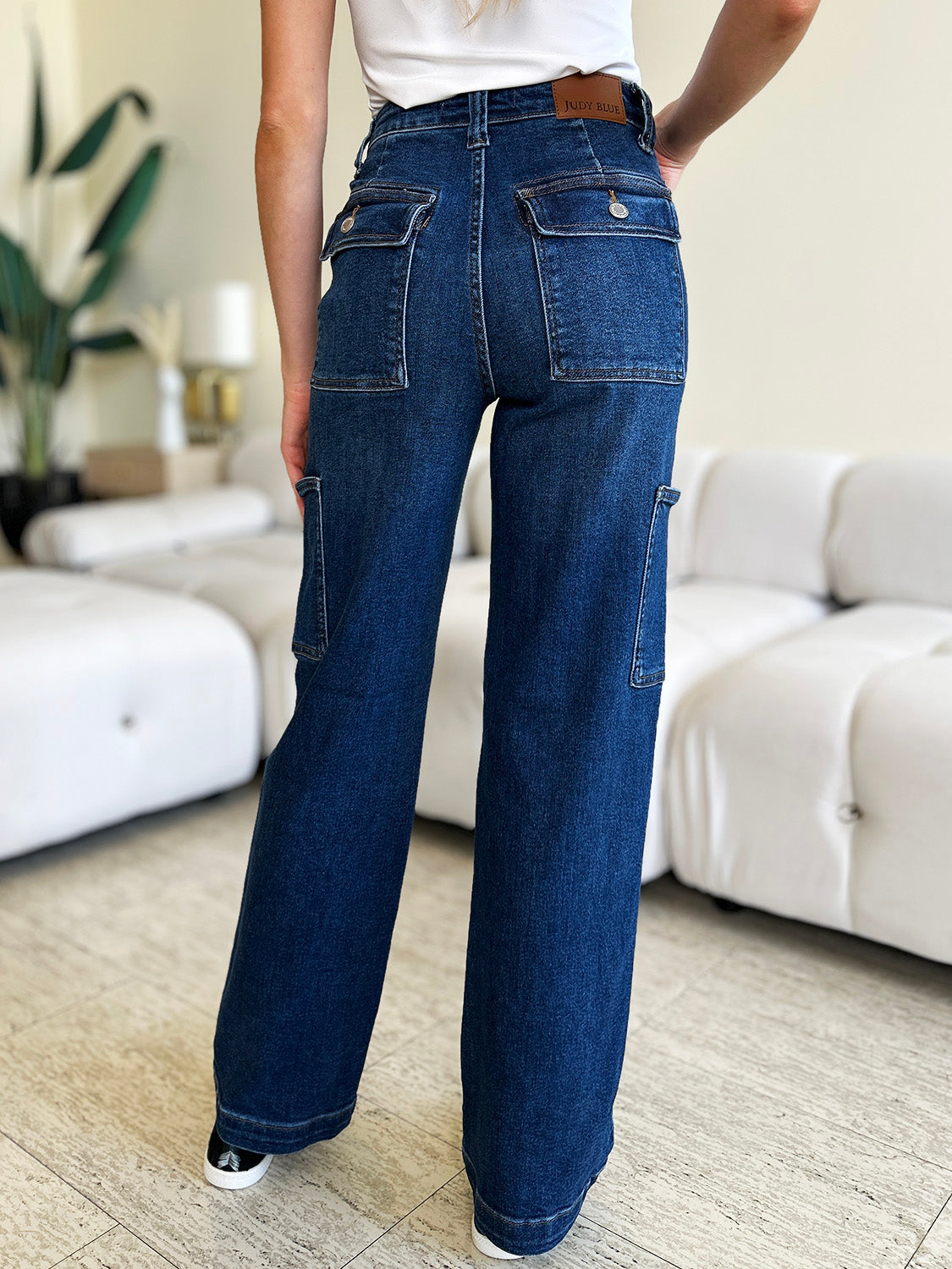 A person wearing Judy Blue Full Size High Waist Straight Cargo Jeans made of durable denim fabric and a white top walks on a wooden floor, with a white sofa and green plant in the background.