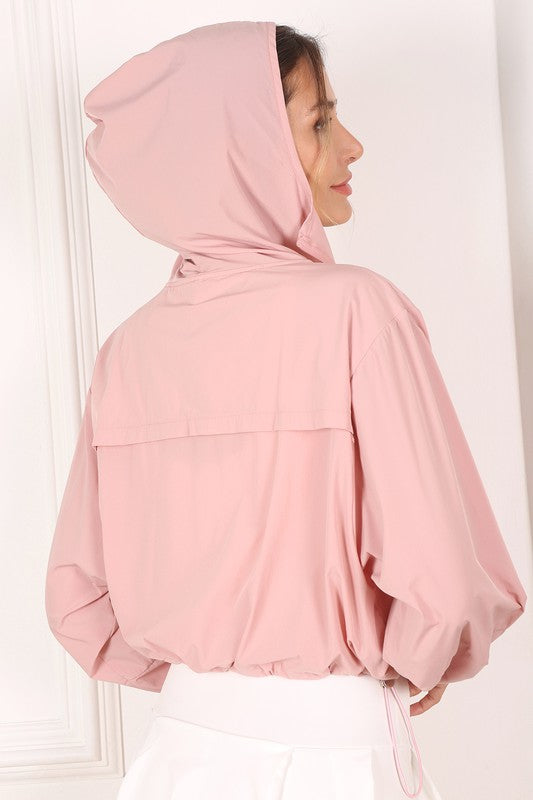 A woman wearing a pink wind breaker over a white top stands against a white wall.