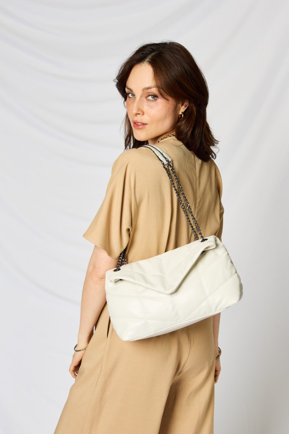 A person holding a stylish SHOMICO PU Leather Chain Handbag over their shoulder against a white background. They are dressed in a beige outfit and looking at the camera, embodying sustainable fashion.