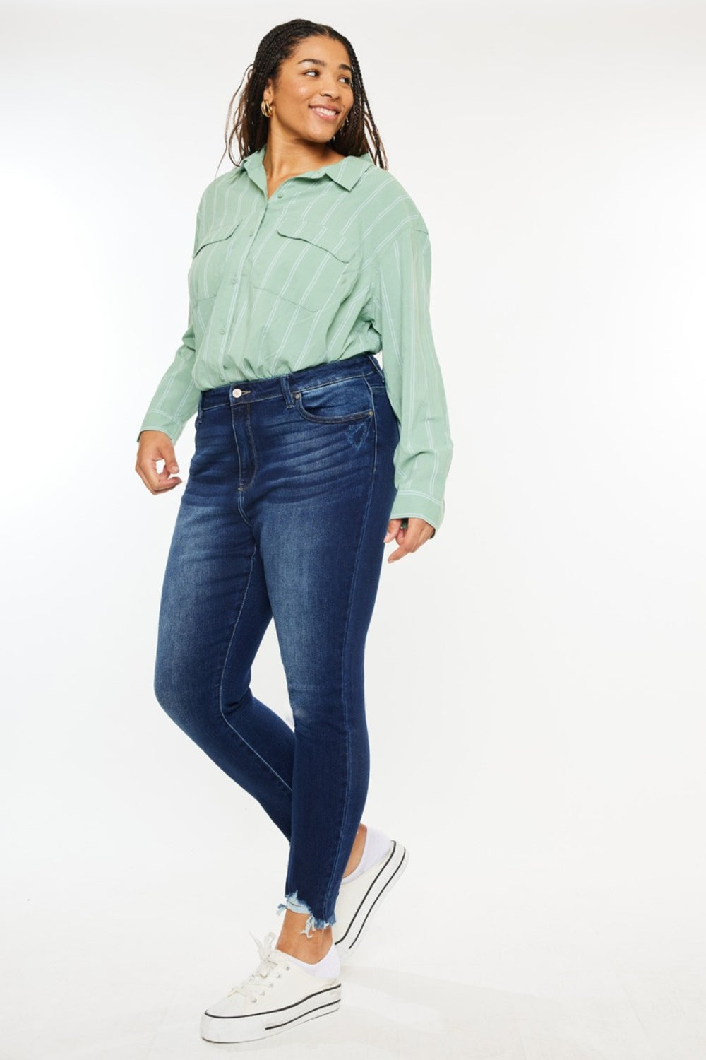 A person wearing a green striped shirt, Kancan Full Size High Rise Frayed Ankle Skinny Jeans, and white sneakers stands smiling against a plain background.