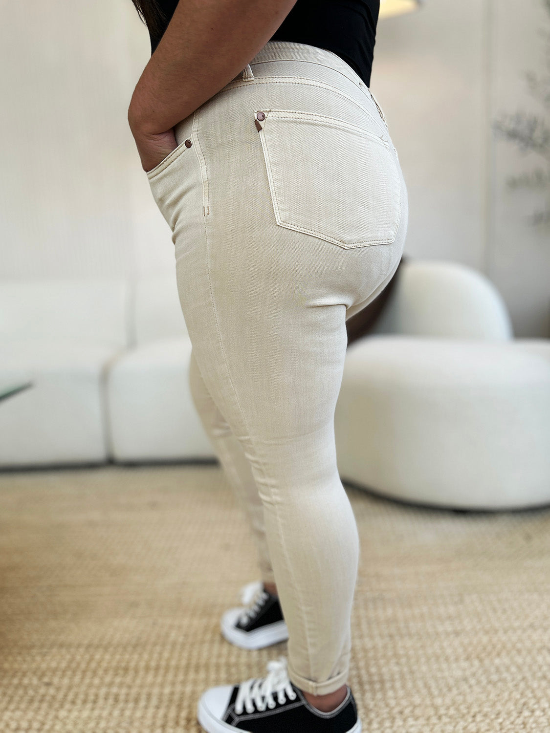 A person wearing Judy Blue Full Size Garment Dyed Tummy Control Skinny Jeans, a white tucked-in top, and white sneakers stands indoors on a woven rug, with one hand in a pocket.