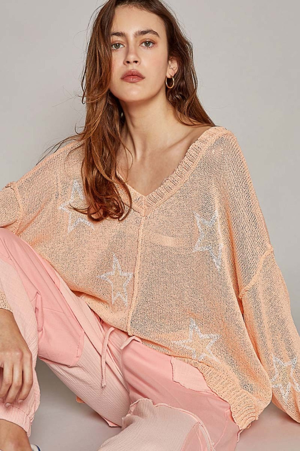 A woman with long hair is sitting down, wearing a fashionable POL V-Neck Long Sleeve Star Print Knit Top in a peach, loose-knit design. The top is paired perfectly with matching peach pants. She looks directly at the camera with a neutral expression.