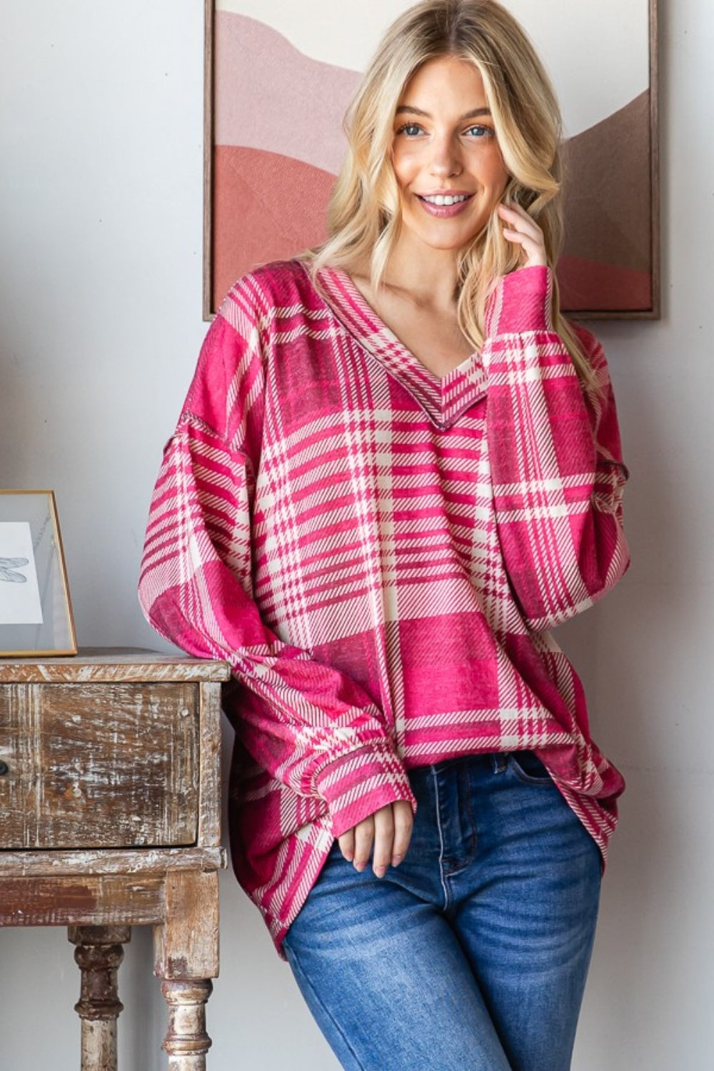 A person with long blond hair smiles while wearing the Heimish Plaid Drop Shoulder V-Neck T-Shirt in red and white and a pair of blue jeans, leaning against a wooden table with framed pictures in the background.