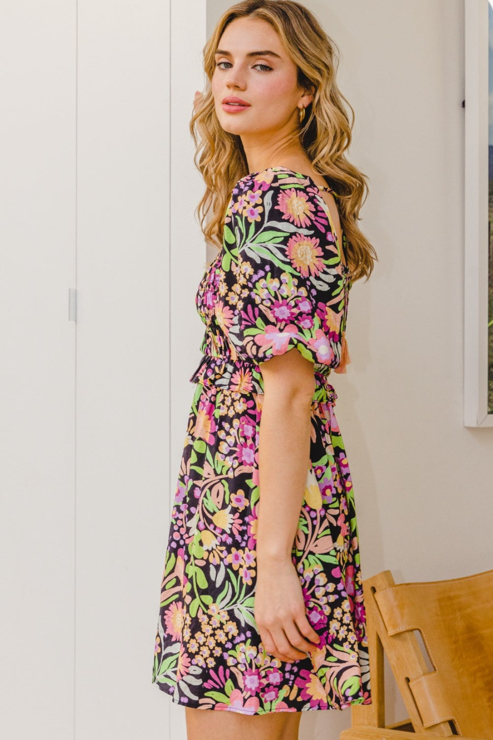 A person stands wearing the ODDI Full Size Floral Tie-Back Mini Dress, a feminine and flirty black floral design with puffy sleeves, perfect for a summer wedding.