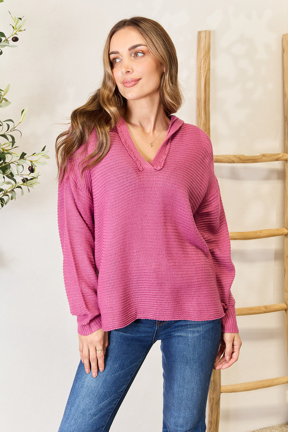 A woman in a stylish Shiny Ribbed Fringed V-Neck Hooded Sweater and jeans stands beside a wooden ladder and a plant.