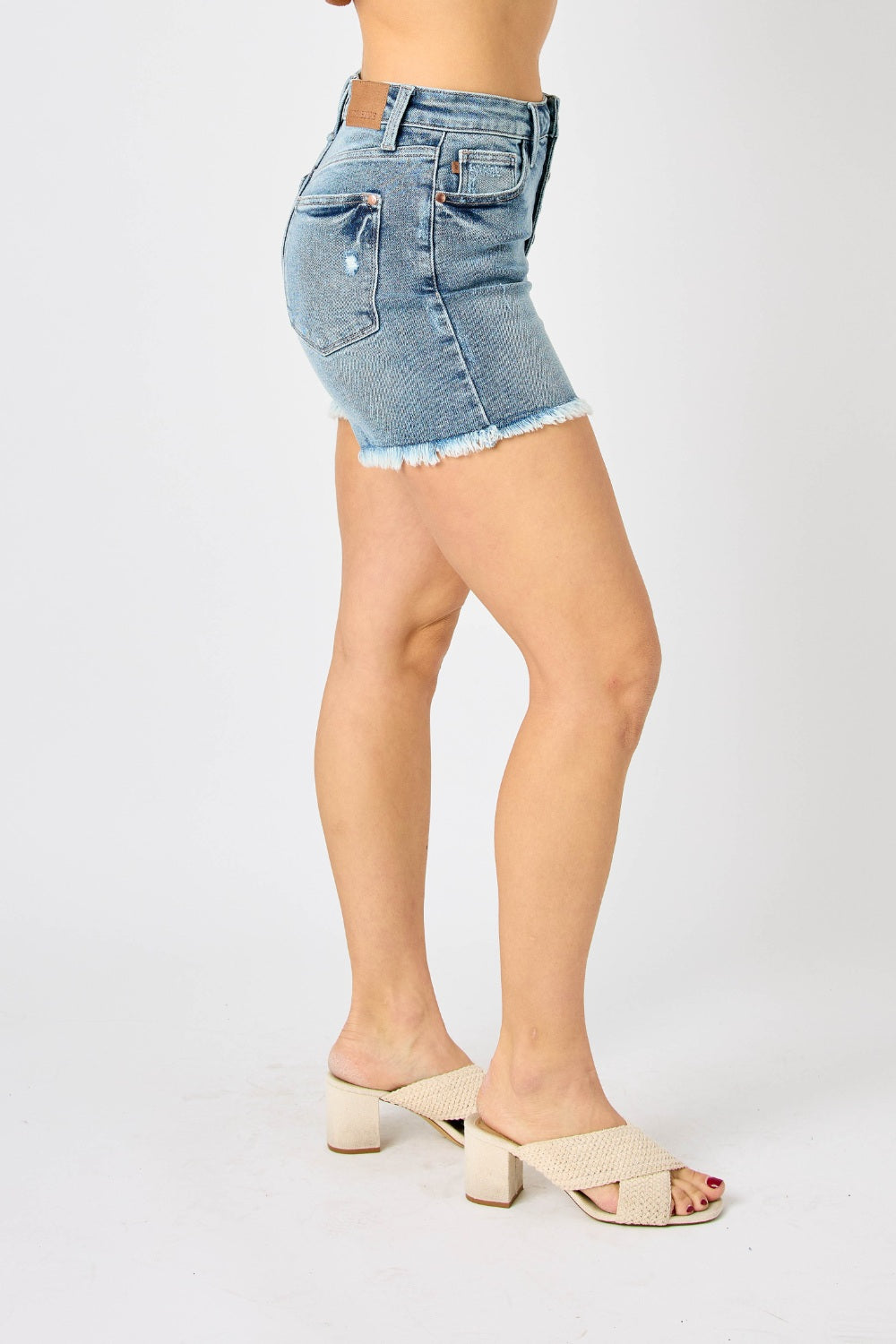 Wearing the Judy Blue Full Size Button Fly Raw Hem Denim Shorts and a white t-shirt, a person stands smiling in sandals against a plain background.