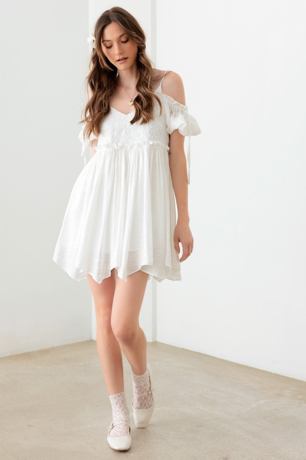 A woman poses in a boho-inspired, embroidered floral shirred cold shoulder mini dress by Mustard Seed. She accessorizes with a gold necklace and maintains a neutral expression against the backdrop of a plain white wall.