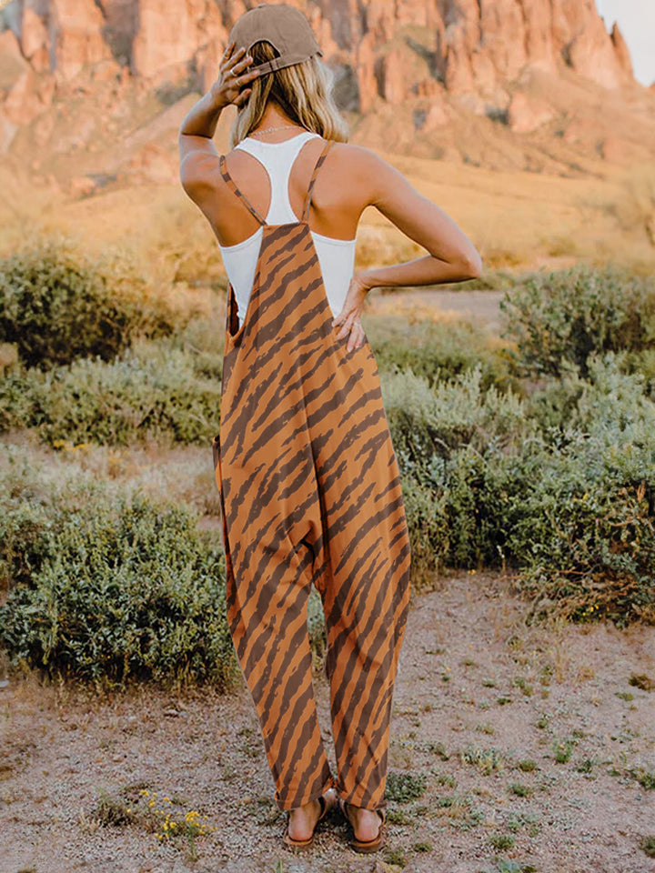 A person wearing a casual white V-neck tank top and a Full Size Printed V-Neck Sleeveless Jumpsuit with a brown animal print zebra-stripe pattern stands outdoors with one hand on their hat and the other in their pocket, set against a rocky landscape backdrop.