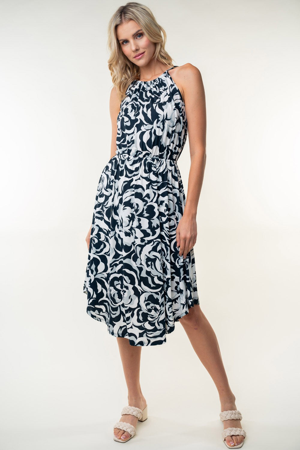 The person wearing a White Birch Tied Ruched Floral Sleeveless Knee Length Dress, featuring a black and white floral pattern, stands and smiles against a neutral background.