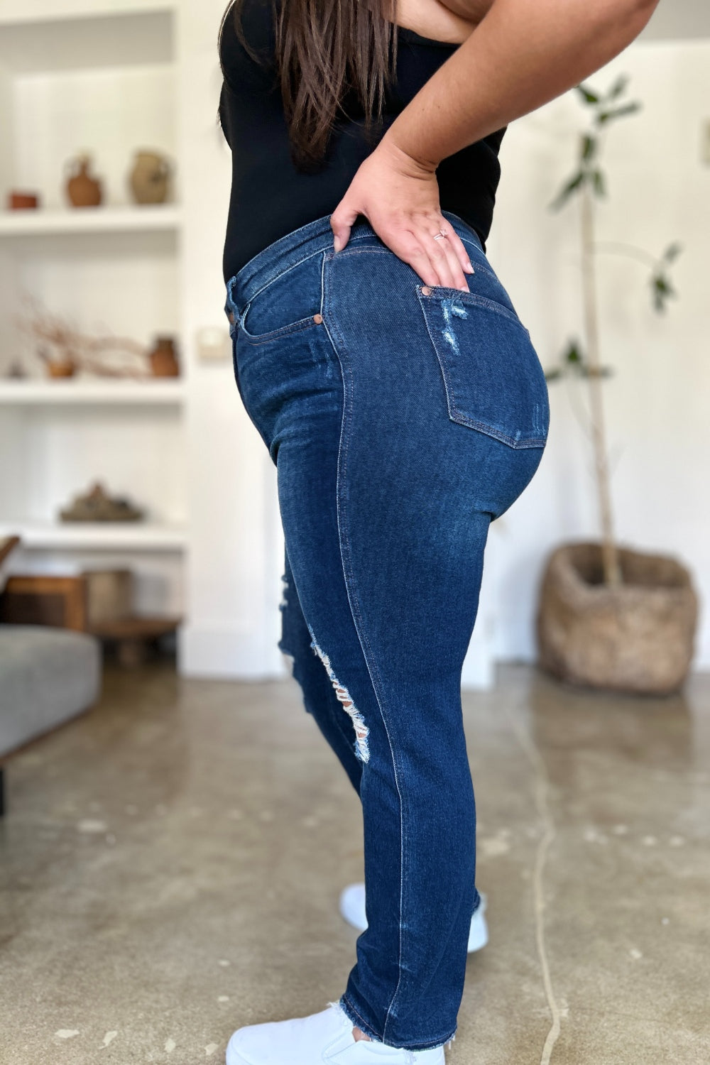 A woman in a black sleeveless top and Judy Blue Full Size High Waist Rigid Magic Heavy Destroy Straight Jeans stands indoors with her hands in her pockets. The background features a plant and shelves adorned with decor items.