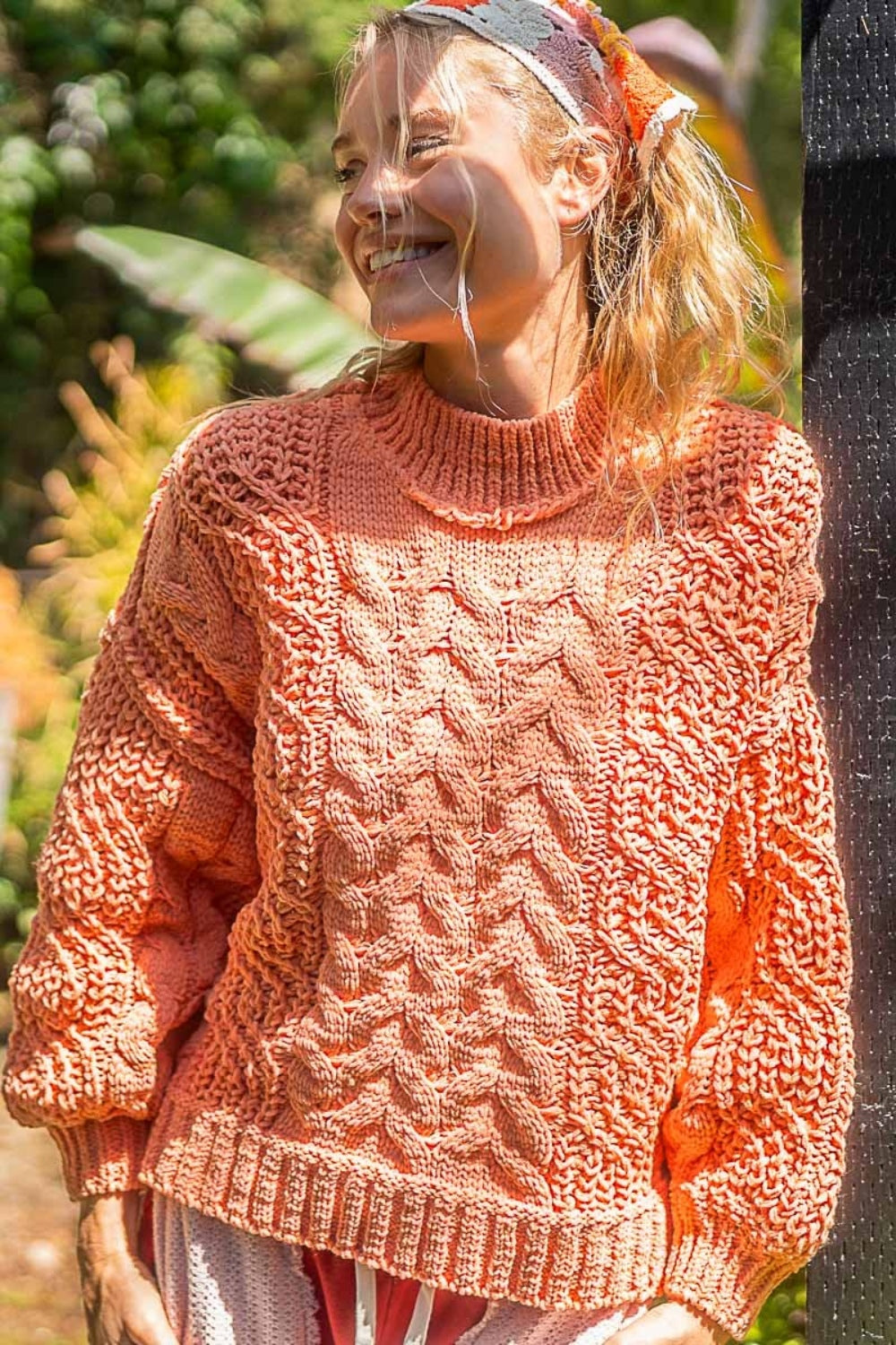 A person standing outdoors, smiling warmly, in a peach POL Mock Neck Cable Knit Sweater and a headband.