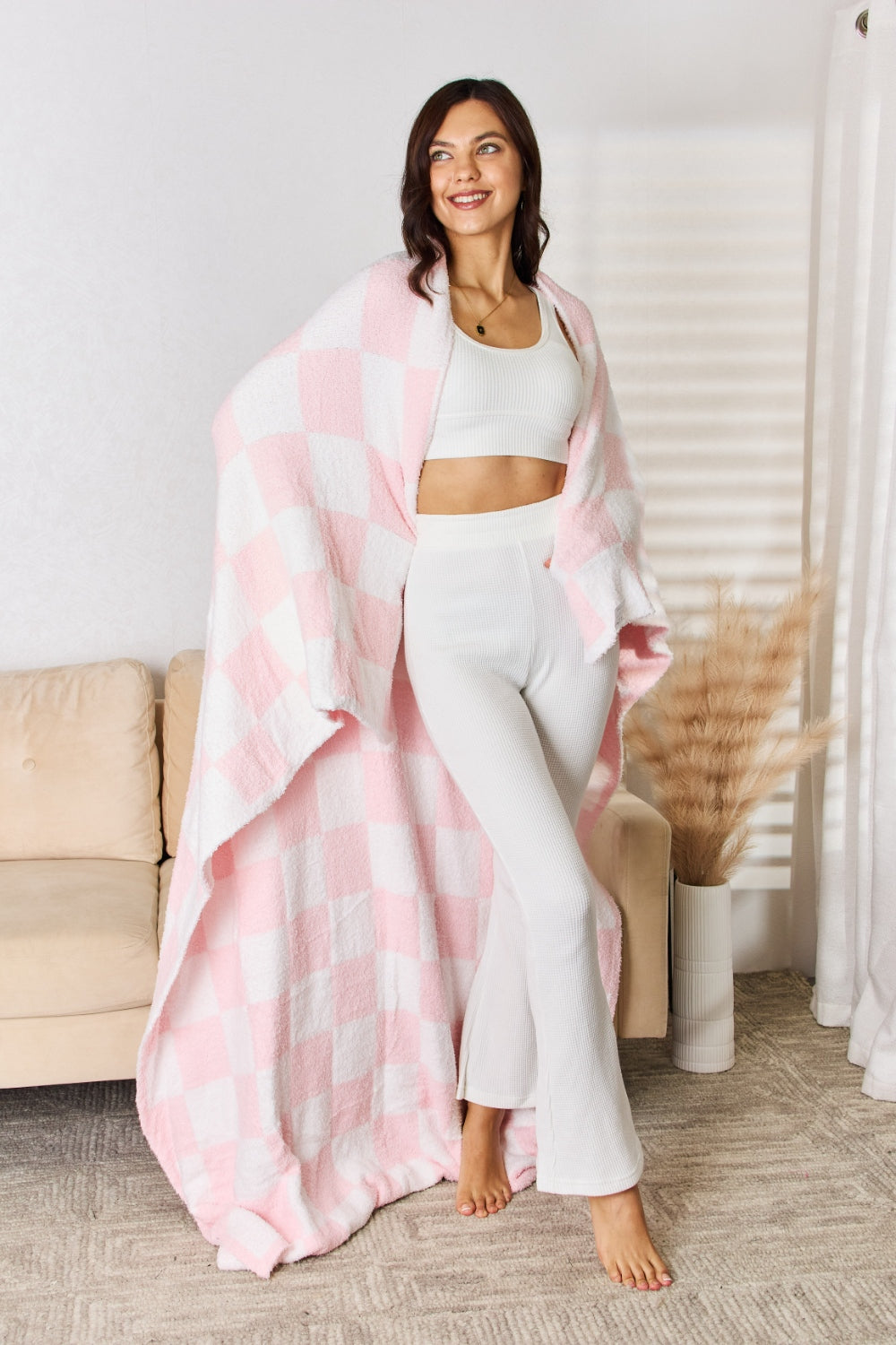 A person stands holding a large Cuddley Checkered Decorative Throw Blanket, smiling, in a room with a sofa and a potted plant. The blanket features a black and white checkerboard pattern, is made from 100% polyester, and imported for quality.