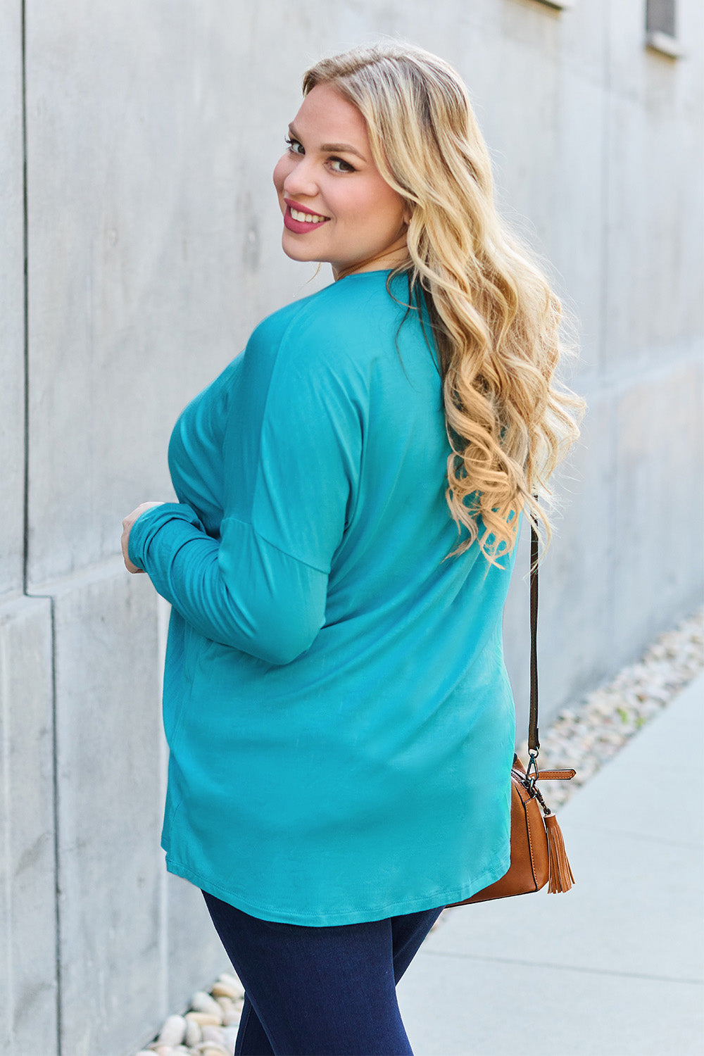 A woman with shoulder-length hair wearing a Basic Bae Full Size Round Neck Dropped Shoulder T-Shirt in green and a pair of jeans, exuding a basic style, stands against a concrete wall. She's holding an imported brown woven backpack on her shoulder while touching her hair with her hand.