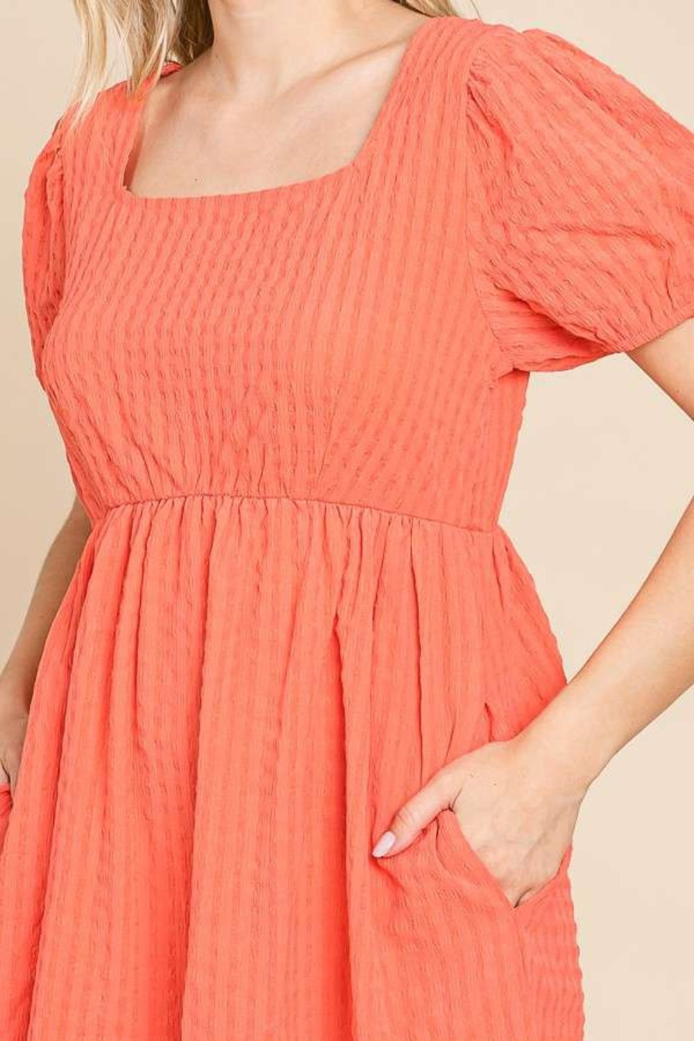 A woman is wearing a Culture Code Textured Square Neck Short Sleeve Dress: it's a stylish, short-sleeved, square-necked, orange dress with puffed sleeves. She is smiling and has one hand in her pocket.