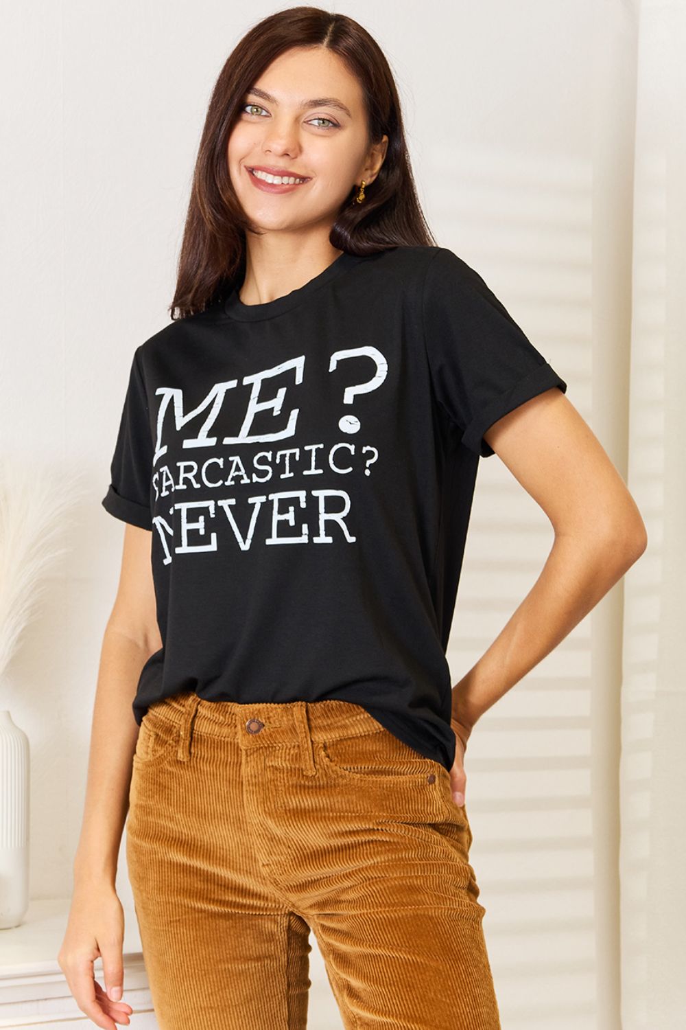 A woman with long dark hair smiles while wearing a trendy black Simply Love Letter Graphic Round Neck T-Shirt featuring the text "ME? Sarcastic? NEVER" and brown corduroy pants. She stands in a room with white walls and decor.