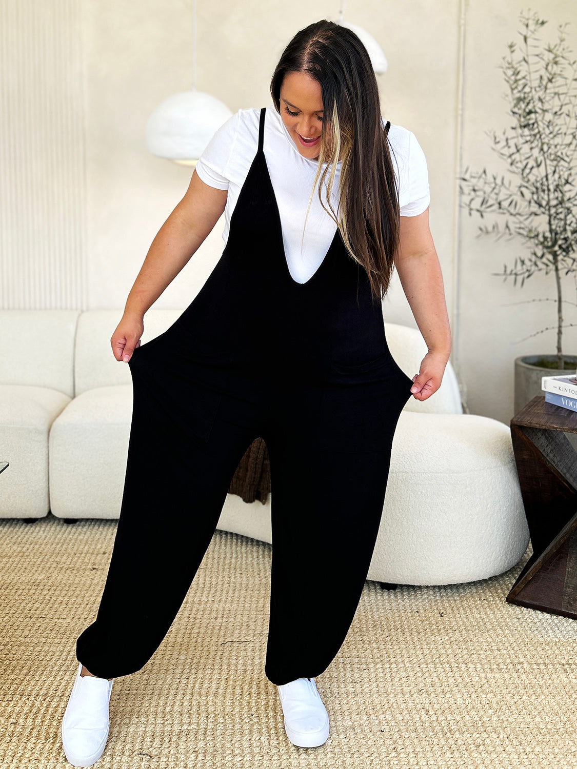 A person wearing the Culture Code Full Size Plunge Sleeveless Jumpsuit with Pockets stands indoors on a beige carpet near a white couch, smiling and holding one hand in their pocket.