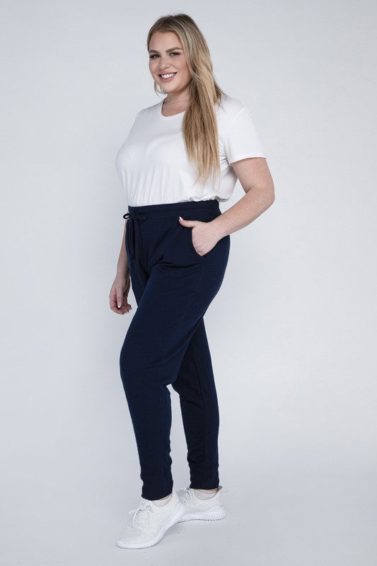 A person stands against a white background, wearing a white t-shirt, grey Plus-Size Jogger Pants with an adjustable waistband, and white sneakers. They have long blonde hair and are posing with one hand on their hip.