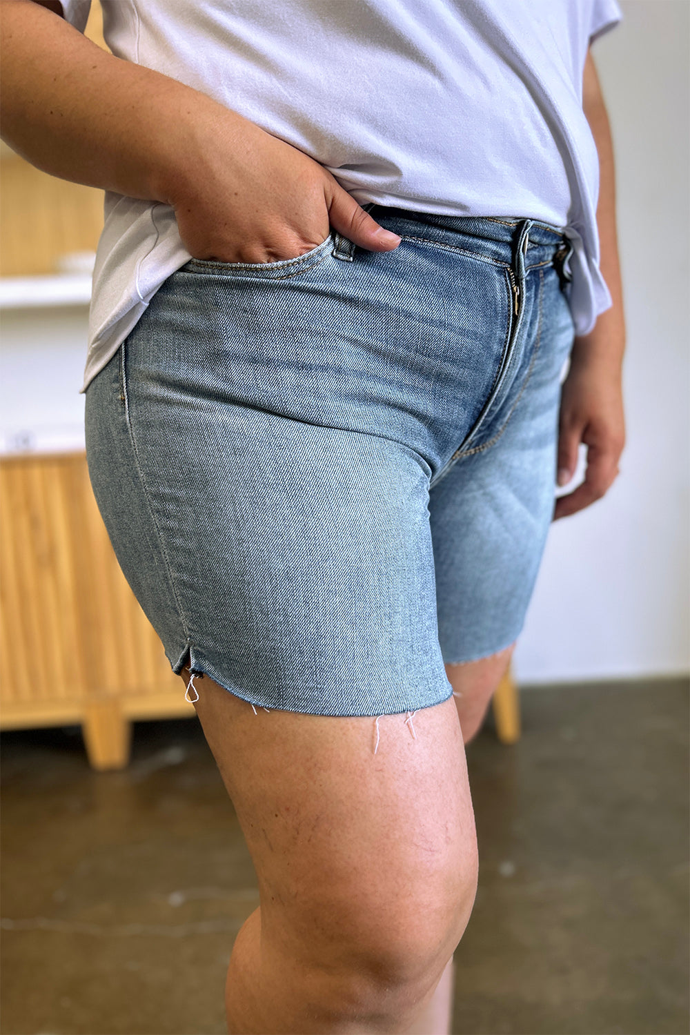 Side view of a person wearing a white shirt and Judy Blue Full Size High Waist Raw Hem Denim Shorts, standing indoors near a wooden cabinet. Summer fashion vibes radiate through the casual ensemble.