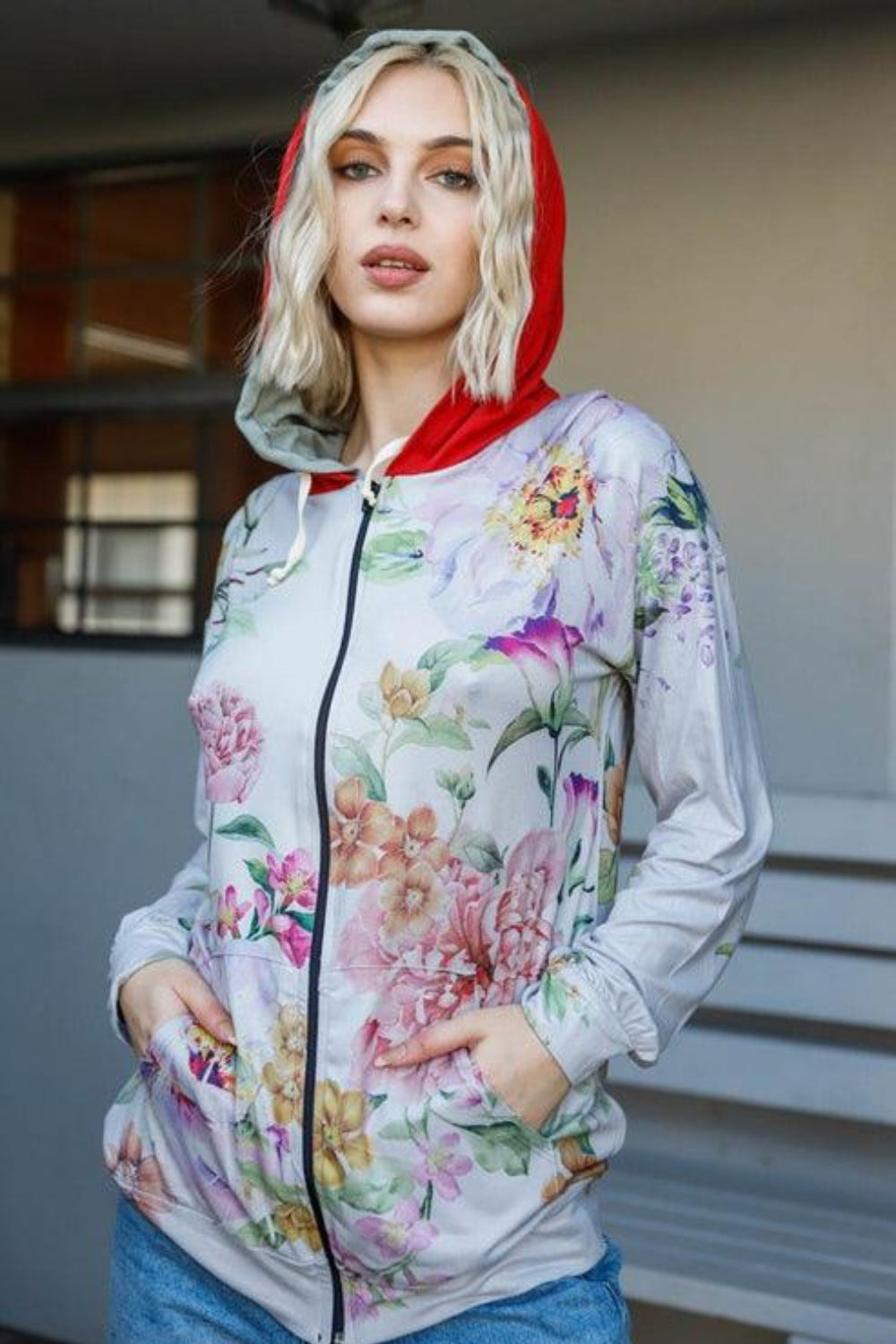 A woman stands outdoors wearing the Blumin Apparel Full Size Floral Zip-Up Hoodie with her hands in the pockets. A leafy green hedge is in the background, adding to the casual chic vibe.