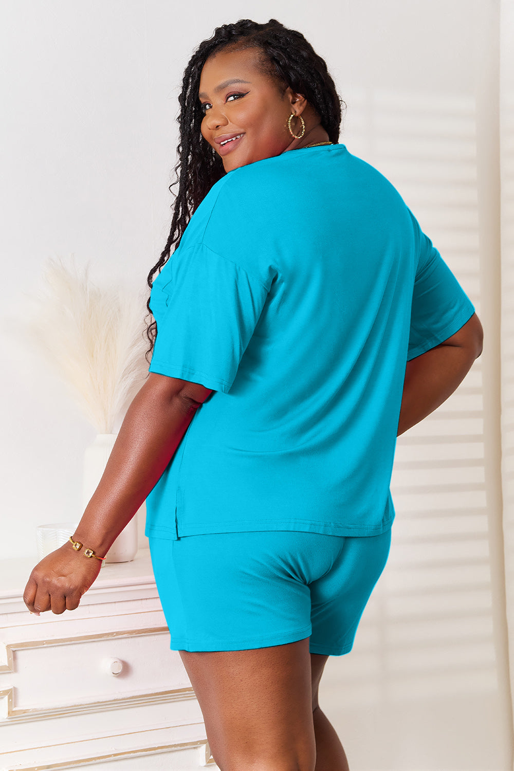 A woman is standing in a brightly lit room, wearing the Basic Bae Full Size Soft Rayon Half Sleeve Top and Shorts Set in a relaxed, stretchy turquoise fabric. She has one hand placed in her pocket and is smiling while looking slightly to the side.