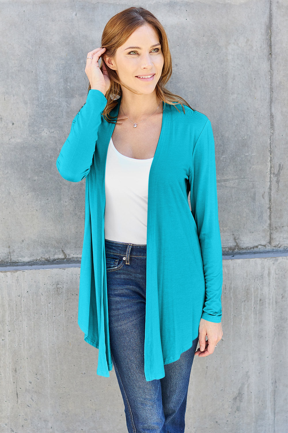 A woman wearing the Basic Bae Full Size Open Front Long Sleeve Cardigan in navy blue, paired with a white top and blue jeans, stands against a concrete wall. She touches her hair and gazes to the side, embodying a chic yet simple style. The slightly stretchy materials of the cardigan ensure comfort while maintaining its stylish simplicity. For easy care, machine wash it cold.