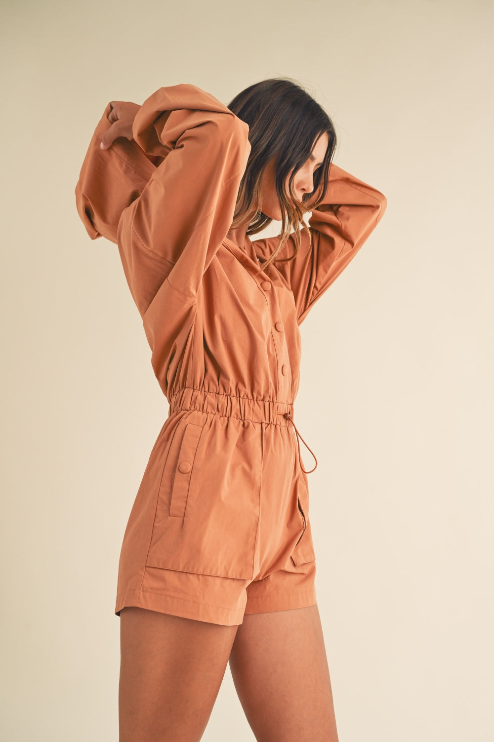 A person wearing the Mable Snap Down Long Sleeve Hooded Romper, featuring a brown color, long sleeves, a snap-down front, and an elastic waistband, is standing against a plain background.