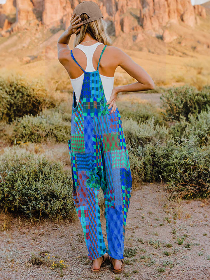 A woman stands outdoors in front of a rocky landscape, wearing the Full Size Printed V-Neck Sleeveless Jumpsuit and a brown brimmed hat. She is smiling and holding the brim of her hat with one hand, exuding the carefree vibe of this vibrant and colorful jumpsuit.