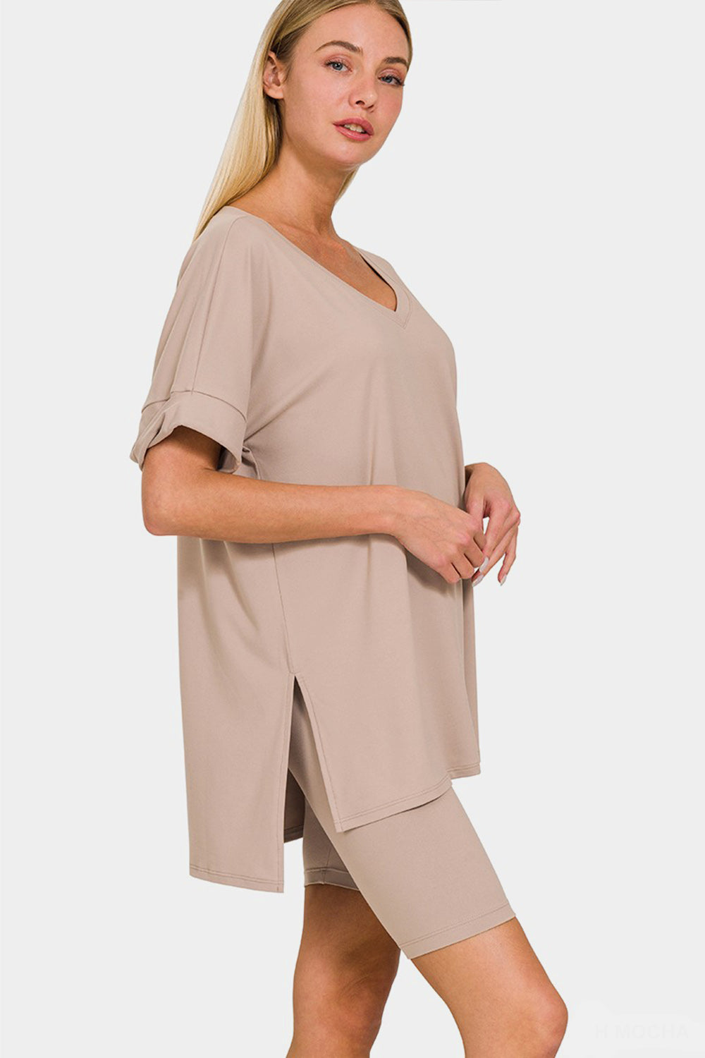 A woman wearing the highly stretchy Zenana Full Size V-Neck Short Sleeve Slit T-Shirt and Shorts Set in beige, featuring a mid-thigh shorts and short sleeve t-shirt combination, stands against a plain background.