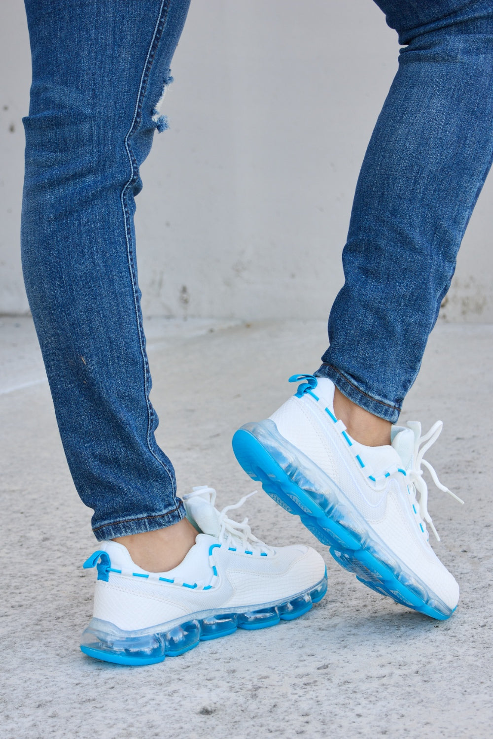 A person wearing ripped jeans and stylish Forever Link Lace-Up Air-Cushioned Athletic Shoes in white with blue accents stands confidently on a concrete surface.