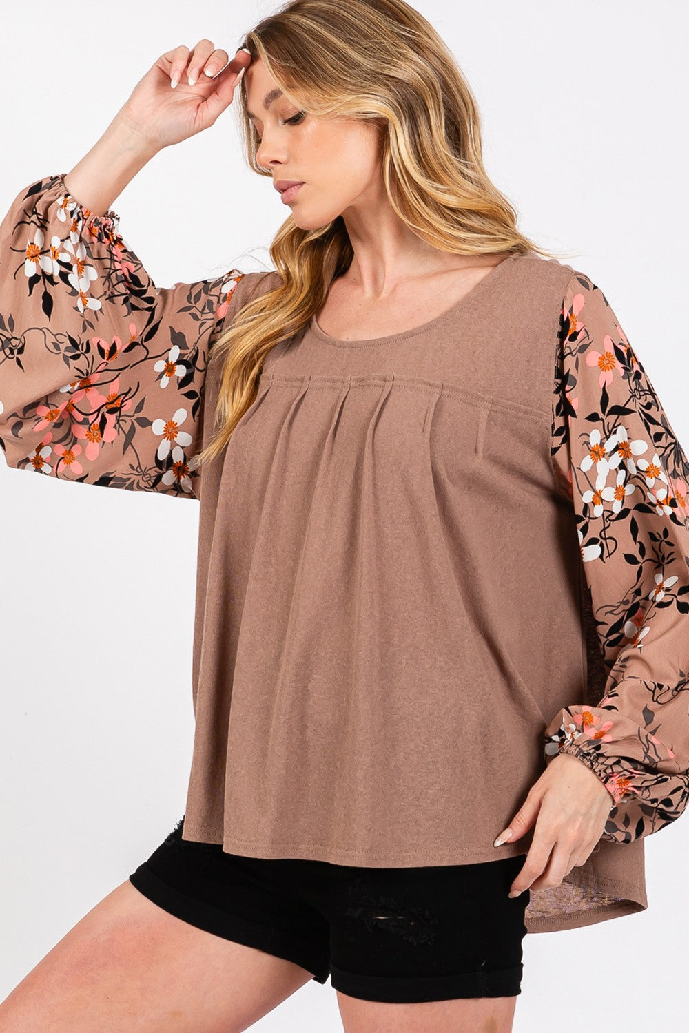 A person is wearing the SAGE + FIG Floral Long Sleeve Front Pleated Detail Blouse, paired with black shorts and a black hat, standing against a plain background.