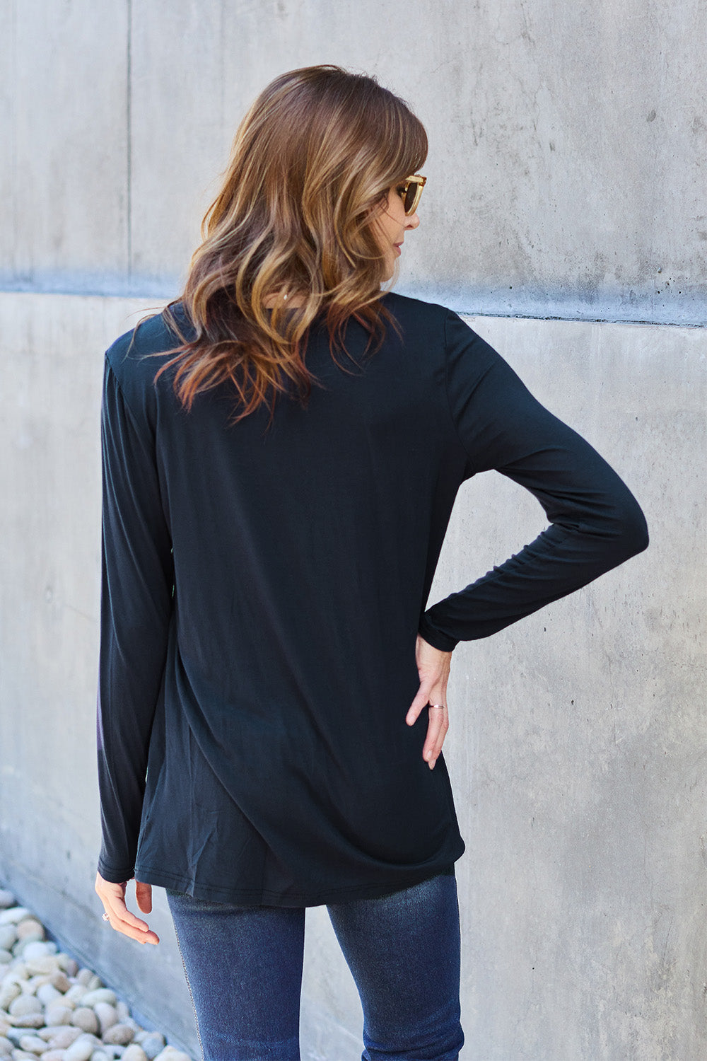 A woman with long hair wearing sunglasses, a Basic Bae Full Size Round Neck Long Sleeve Top in turquoise, and jeans is standing in front of a concrete wall with one hand on her hip.