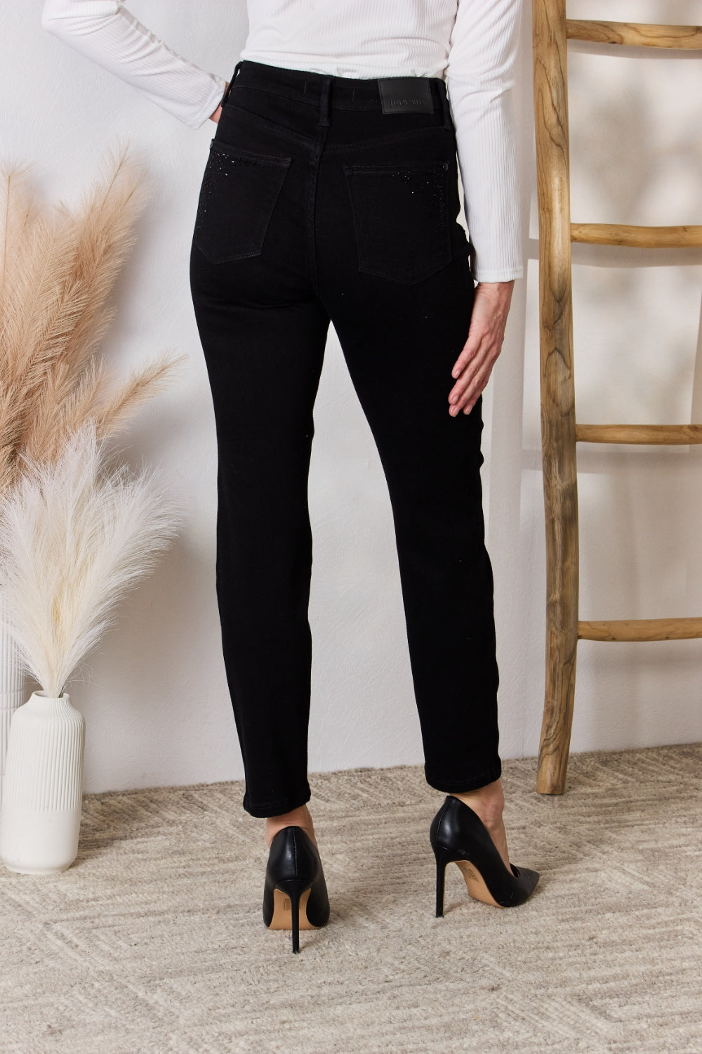 A person wearing slightly stretchy, black Judy Blue Full Size Rhinestone Embellished Slim Jeans and black shoes stands on a textured floor next to a white vase with brown and white ornamental grasses and a wooden ladder.