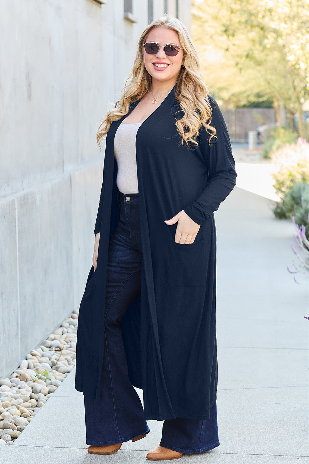 A woman stands against a concrete wall, wearing the Basic Bae Full Size Open Front Long Sleeve Cover Up made from rayon spandex, a white top, blue jeans in her perfect size, and brown shoes with her hands in her pockets. Machine wash cold for best results.