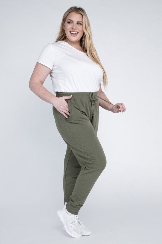 A person stands against a white background, wearing a white t-shirt, grey Plus-Size Jogger Pants with an adjustable waistband, and white sneakers. They have long blonde hair and are posing with one hand on their hip.